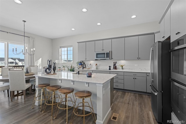 kitchen with appliances with stainless steel finishes, dark wood-type flooring, hanging light fixtures, gray cabinets, and a center island with sink