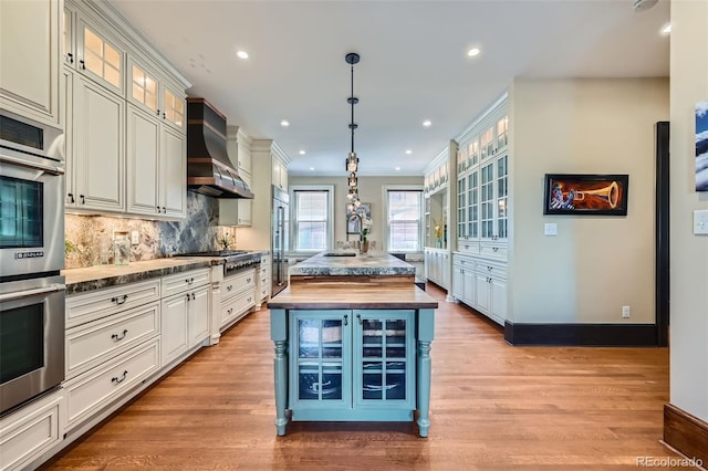 kitchen with appliances with stainless steel finishes, wall chimney exhaust hood, decorative light fixtures, decorative backsplash, and a center island with sink