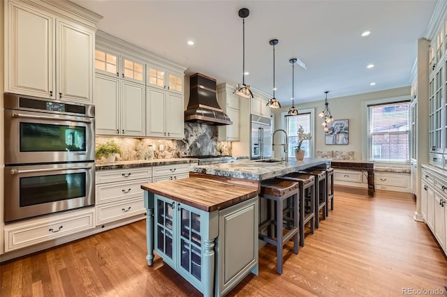 kitchen with an island with sink, tasteful backsplash, custom range hood, wood counters, and appliances with stainless steel finishes