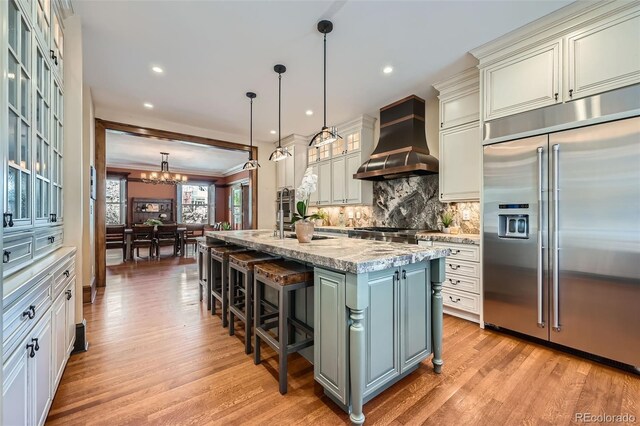 kitchen featuring custom exhaust hood, built in fridge, tasteful backsplash, light stone counters, and a notable chandelier