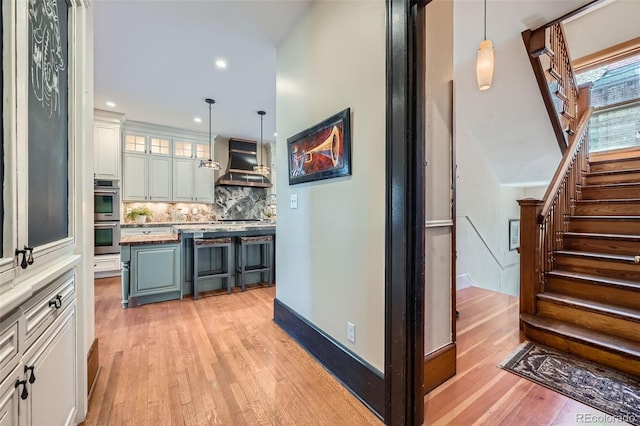 bar with wall chimney range hood, decorative backsplash, stairway, double oven, and light wood-style floors