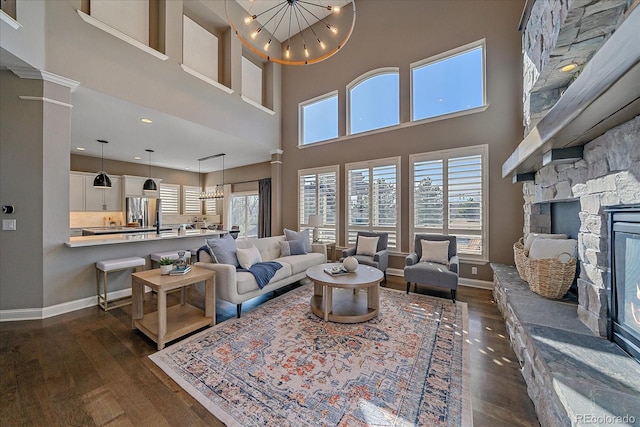 living room with baseboards, a chandelier, decorative columns, a stone fireplace, and dark wood-style flooring