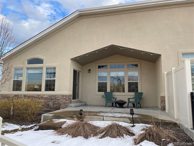 snow covered property entrance featuring stone siding, a patio area, and stucco siding