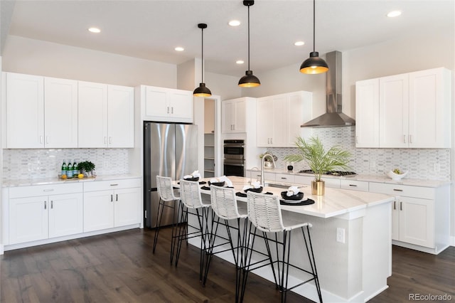 kitchen featuring appliances with stainless steel finishes, hanging light fixtures, an island with sink, wall chimney exhaust hood, and white cabinets