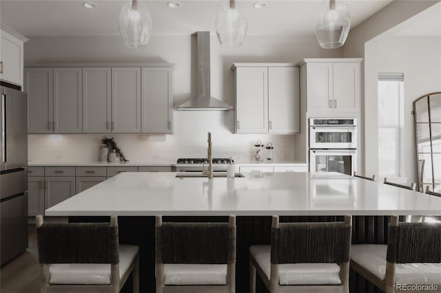 kitchen with hanging light fixtures, a large island with sink, stainless steel refrigerator, double wall oven, and wall chimney range hood