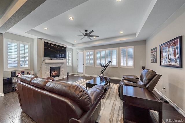 living room with a raised ceiling, a tiled fireplace, ceiling fan, and wood-type flooring