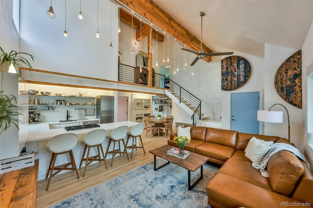 living room with a high ceiling, light hardwood / wood-style floors, sink, ceiling fan, and beam ceiling