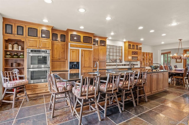 kitchen with a breakfast bar, stainless steel double oven, hanging light fixtures, and an island with sink