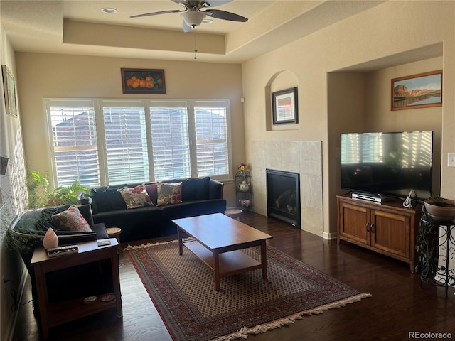 living room with a tiled fireplace, dark wood-type flooring, ceiling fan, and a raised ceiling