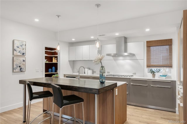 kitchen featuring an island with sink, a kitchen breakfast bar, wall chimney exhaust hood, light wood finished floors, and stainless steel gas cooktop