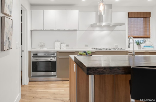 kitchen featuring appliances with stainless steel finishes, wall chimney range hood, light wood-type flooring, white cabinets, and hanging light fixtures