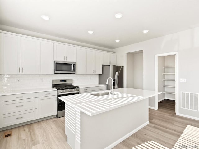 kitchen featuring light wood finished floors, a center island with sink, decorative backsplash, appliances with stainless steel finishes, and light countertops