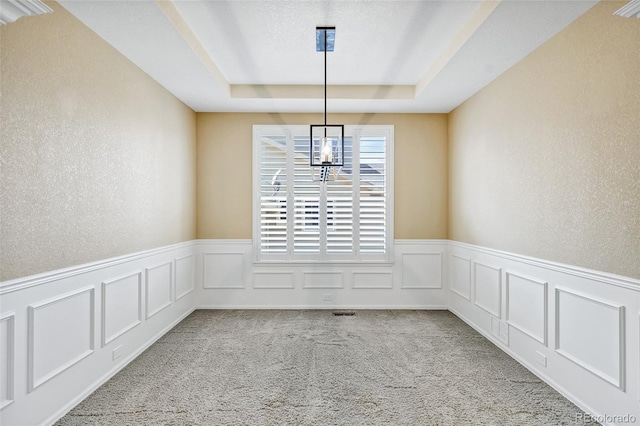unfurnished dining area with a tray ceiling, a decorative wall, carpet floors, a wainscoted wall, and a chandelier