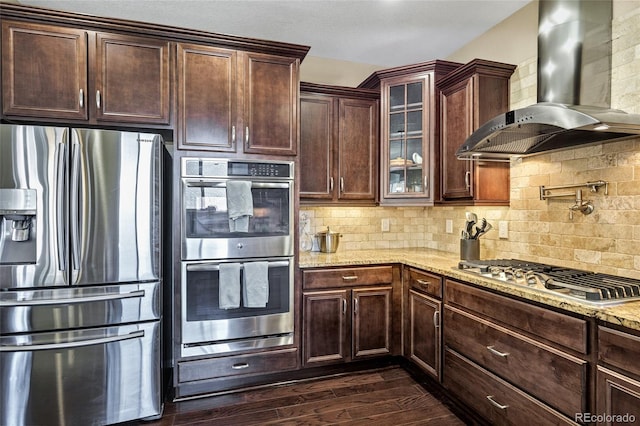 kitchen with appliances with stainless steel finishes, wall chimney range hood, light stone countertops, glass insert cabinets, and dark wood-style flooring
