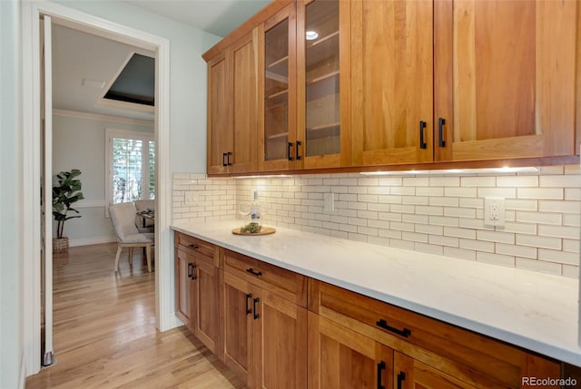 kitchen with glass insert cabinets, ornamental molding, decorative backsplash, light wood-style floors, and brown cabinetry