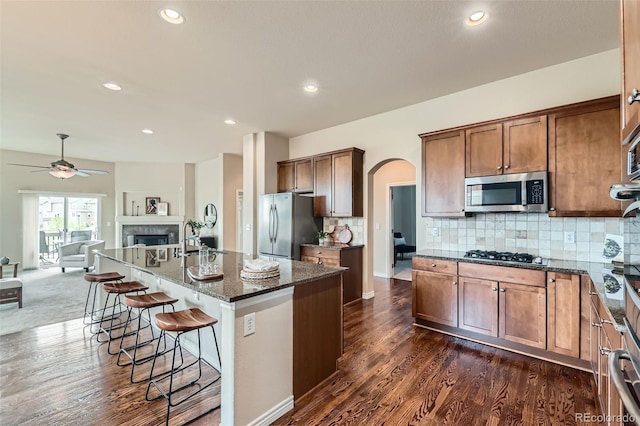 kitchen featuring a breakfast bar area, a kitchen island with sink, appliances with stainless steel finishes, arched walkways, and dark stone countertops