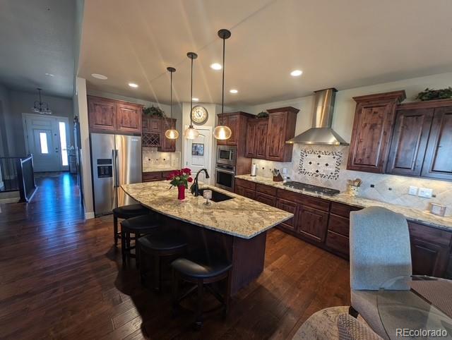 kitchen featuring dark wood finished floors, wall chimney exhaust hood, light stone countertops, stainless steel appliances, and a sink