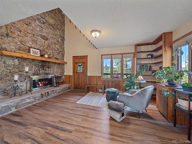 living room featuring a textured ceiling, high vaulted ceiling, wood-type flooring, a stone fireplace, and wooden walls