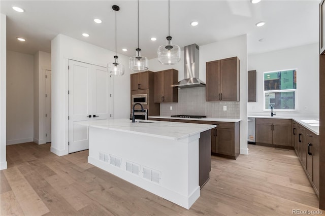 5626 W 142nd Ave, Broomfield, CO 80020 - kitchen with wall chimney exhaust hood, appliances with stainless steel finishes, an island with sink, tasteful backsplash, and light wood-style floors