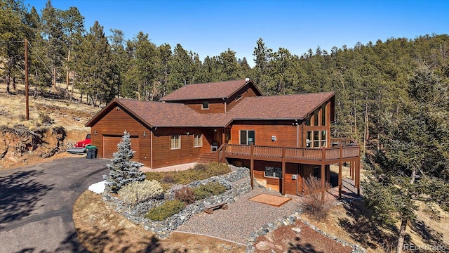 chalet / cabin featuring a garage, a deck, aphalt driveway, and a wooded view