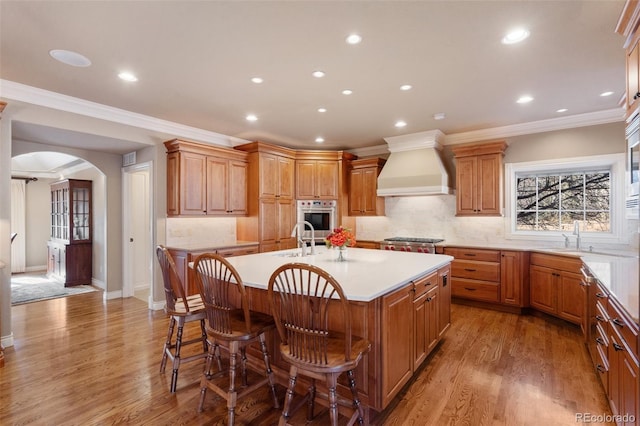 kitchen with premium range hood, stainless steel appliances, a kitchen island with sink, wood-type flooring, and a breakfast bar area