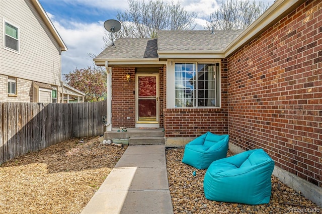 view of exterior entry with brick siding, roof with shingles, and fence