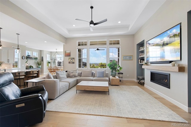 living room featuring a tray ceiling, light wood-style floors, a ceiling fan, and a glass covered fireplace