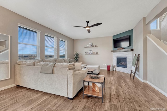 living area featuring a glass covered fireplace, ceiling fan, baseboards, and wood finished floors