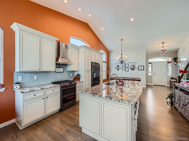 kitchen featuring wall chimney range hood, an inviting chandelier, double oven range, an island with sink, and black refrigerator with ice dispenser