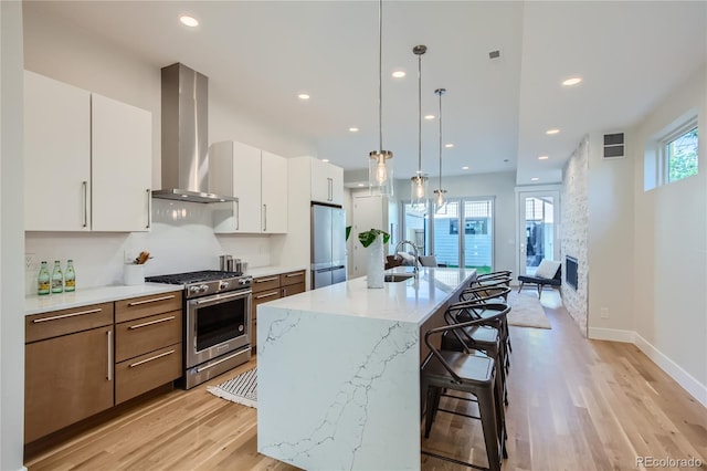 kitchen featuring hanging light fixtures, wall chimney exhaust hood, an island with sink, appliances with stainless steel finishes, and white cabinetry