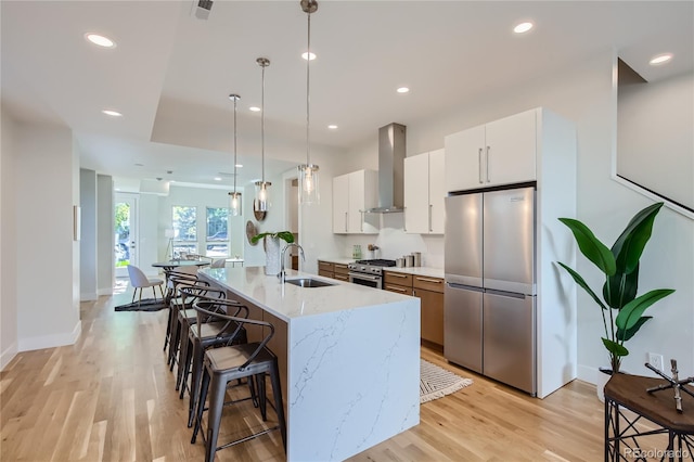 kitchen with stainless steel appliances, a kitchen island with sink, wall chimney range hood, decorative light fixtures, and white cabinetry