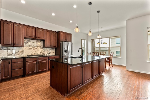 kitchen featuring sink, stainless steel fridge with ice dispenser, pendant lighting, dark stone counters, and a kitchen island with sink