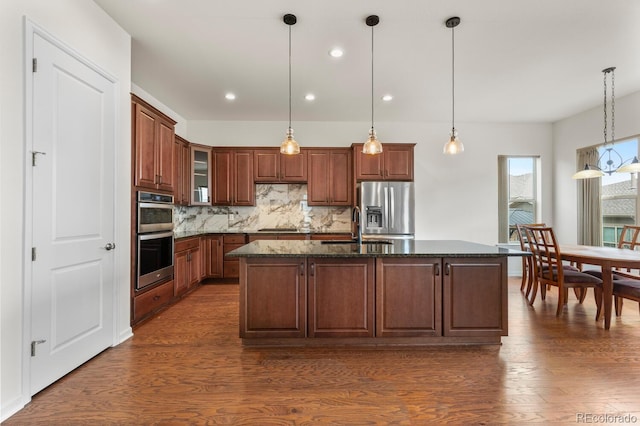 kitchen featuring appliances with stainless steel finishes, decorative light fixtures, a center island with sink, and dark stone countertops