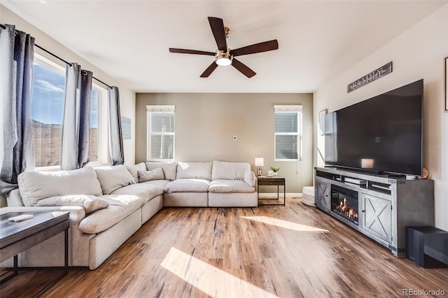 living area featuring a glass covered fireplace, ceiling fan, baseboards, and wood finished floors