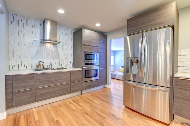 kitchen with wall chimney range hood, light wood-type flooring, tasteful backsplash, and stainless steel appliances