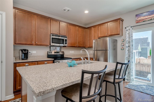 kitchen with a kitchen breakfast bar, appliances with stainless steel finishes, brown cabinets, an island with sink, and dark wood finished floors