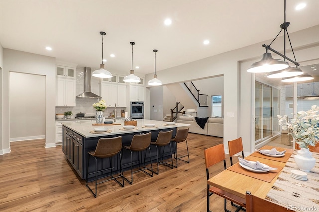 kitchen with oven, wall chimney exhaust hood, pendant lighting, white cabinetry, and light hardwood / wood-style floors