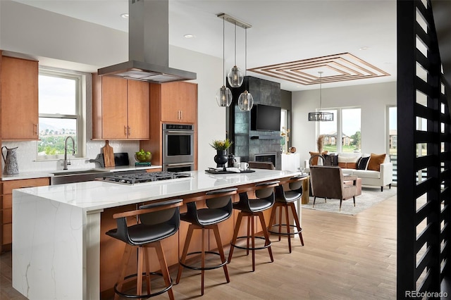 kitchen with light hardwood / wood-style floors, stainless steel appliances, a tiled fireplace, hanging light fixtures, and island exhaust hood