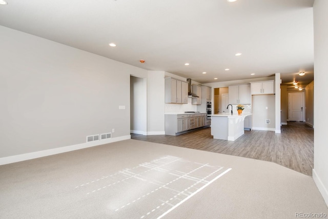 kitchen featuring light countertops, a kitchen island with sink, wall chimney exhaust hood, baseboards, and black cooktop
