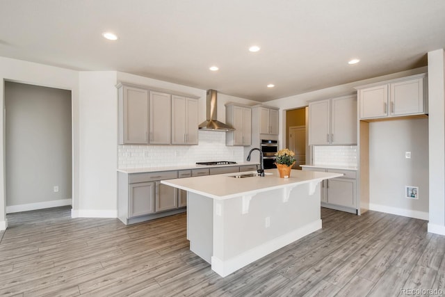 kitchen featuring a sink, light wood-type flooring, wall chimney exhaust hood, a kitchen island with sink, and appliances with stainless steel finishes