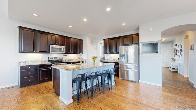 kitchen featuring stainless steel appliances, an island with sink, light stone countertops, and light wood-type flooring