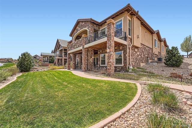 rear view of house with a lawn, a balcony, stone siding, and stucco siding