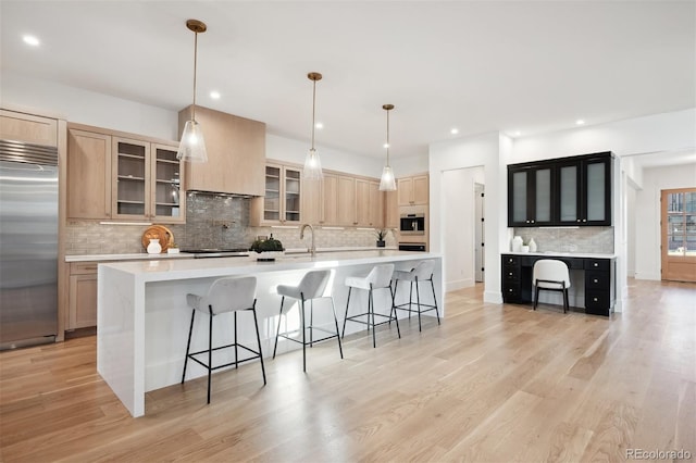 kitchen with light brown cabinets, built in appliances, decorative light fixtures, light hardwood / wood-style flooring, and a large island with sink