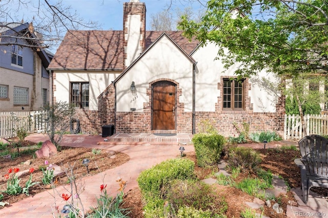 tudor-style house featuring brick siding, fence, a chimney, and stucco siding