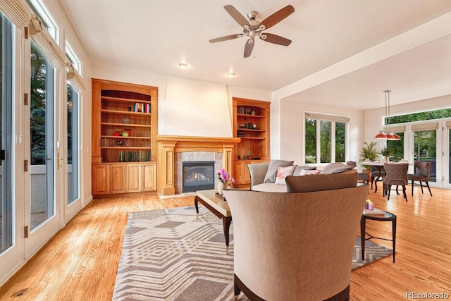 living room featuring a tiled fireplace, ceiling fan, light wood-type flooring, and built in shelves