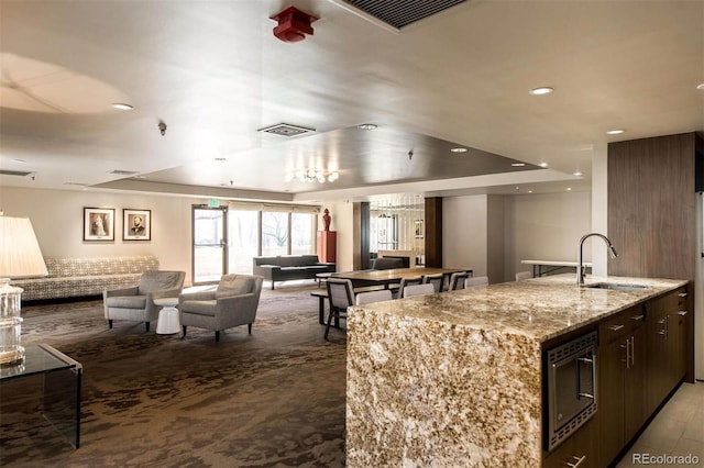 kitchen with stainless steel microwave, sink, dark brown cabinetry, a kitchen island with sink, and light stone counters