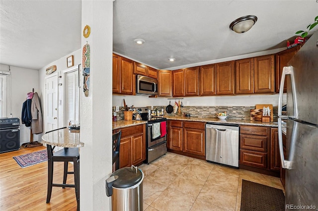 kitchen featuring brown cabinetry, light stone counters, backsplash, light tile patterned floors, and stainless steel appliances