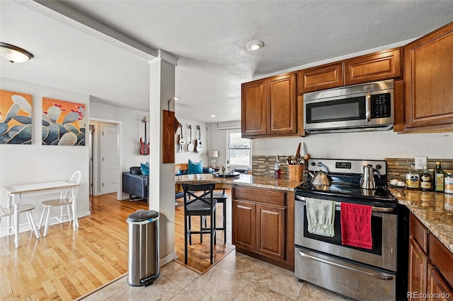 kitchen with a textured ceiling, brown cabinets, stone countertops, stainless steel appliances, and light wood-style flooring