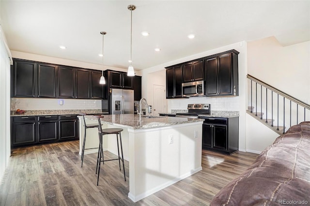 kitchen featuring light wood finished floors, a center island with sink, appliances with stainless steel finishes, light stone counters, and a breakfast bar area