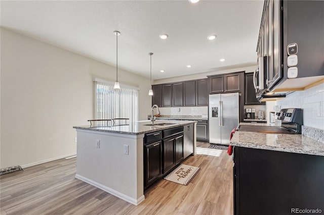 kitchen with light wood finished floors, appliances with stainless steel finishes, a sink, an island with sink, and light stone countertops